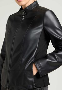 Zwarte aansluitende jas met leren voorkant en stoffen zijkanten, voorzien van een hoge kraag, ritsluiting en zijzakken. Effen textuur, slank ontwerp.