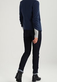 Blazer de tela suave en azul marino con mangas fruncidas, combinado con jeans oscuros y botas negras de tobillo. Los puños presentan una textura de punto gris claro.