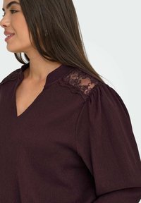 Haut bordeaux avec un décolleté en V, tissu texturé et accents en dentelle sur les épaules. Présente des manches bouffantes et une coupe décontractée.