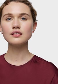 Top bordeaux con una texture liscia, scollatura tonda e una leggera lucentezza. Presenta orecchini argentati con accenti rossi. Pelle chiara e occhi blu.