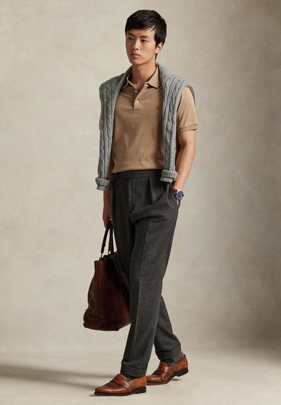 Beige polo-skjorte, grå kabelstrikket genser over skuldrene, grå ullbukser, brune skinnsko, bærer en brun skinnveske.