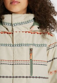 Beige Jacke mit geometrischen Mustern in Grün, Orange und Blau. Sie verfügt über eine Reißverschlusstasche und einen hohen Kragen für zusätzliche Wärme.