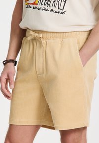 Beige corduroy-shortsit, joissa on joustava vyötärö ja naru, sivutaskut sekä rakenne, jossa on viistoja kohoja.