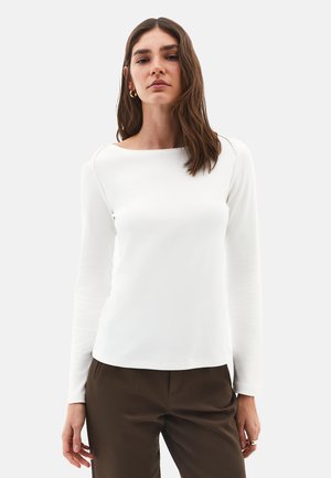 Femme portant un haut blanc à manches longues et un pantalon marron, avec des cheveux bruns lâches et des boucles d'oreilles en or, se tenant devant un fond uni.