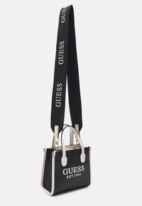 Sac à main noir avec des accents crème, présentant un design structuré, deux poignées supérieures et une longue bandoulière marquée "GUESS" en blanc.