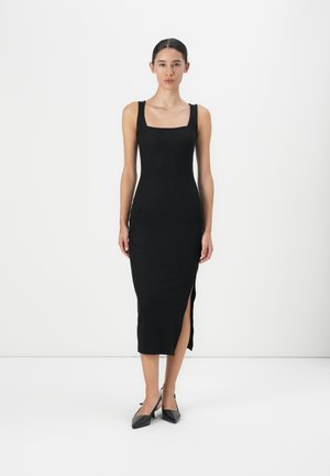 Robe midi sans manches noire avec un décolleté carré, texture lisse, fente sur le côté et coupe ajustée, associée à des escarpins noirs à bout pointu.