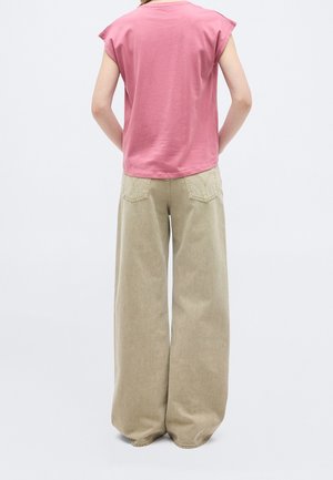 Personne portant un haut ample rose à manches courtes et un pantalon beige large, debout avec les mains derrière le dos sur un fond blanc.