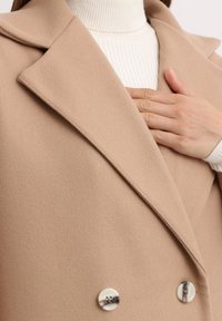 Abrigo beige de doble botonadura con una textura suave, cuello con muesca y botones brillantes, llevado sobre un suéter de cuello alto blanco con canalé.
