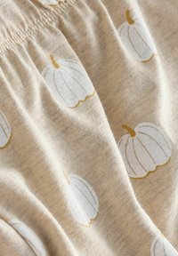 Beige tyg med en mjuk textur som har vita pumpamönster konturerade i guld. Samlad resår upptill skapar en subtil volangeffekt.
