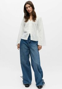 Kvinne med langt brunt hår iført hvit bluse med blomsterbroderi og vide blå jeans med knyttede kanter, står foran en hvit bakgrunn.