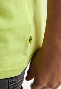 Camiseta de algodón de un brillante color amarillo que muestra la costura de la manga y una pequeña etiqueta de logo negro. Una mano agarra la tela cerca del dobladillo de la camiseta y la piel texturizada.