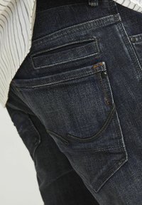 Jeans din denim de culoare albastră închisă, cu o vedere detaliată a buzunarelor din spate. Au cusături în contrast, o finisare texturată și mai multe modele de buzunare.