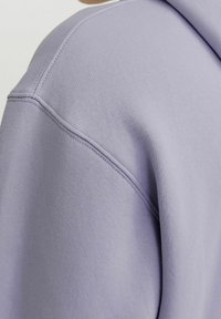 Šviesiai violetinis hoodie, pagamintas iš minkštos medžiagos, turi apvalų iškirpimą ir matomus siūlus peties siūlėse. Lygi tekstūra ir laisvas siluetas.