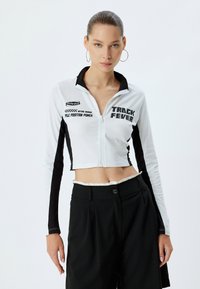 Giacca cropped bianca e nera con zip e maniche lunghe, caratterizzata da stampe grafiche e un design del colletto sportivo. Texture della superficie liscia.