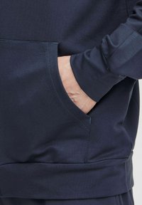 Námornícky modrý sweatshirt s vreckom na prednej strane, vyrobený z textúrovanej tkaniny. Obsahuje rebrované manžety a uvoľnený strih.