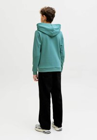 Dreng stående med ryggen mod, iført en teal hættetrøje, sorte bukser og hvide sneakers på en ensartet hvid baggrund.