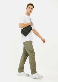 Sac bandoulière noir en nylon avec fermeture éclair, détail de logo, et sangle réglable. Associé à un T-shirt blanc et un pantalon vert olive. Baskets blanches.