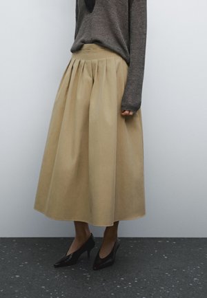 Personne portant une jupe midi plissée beige, un pull à manches longues gris foncé et des chaussures noires à talons hauts, debout sur un sol sombre tacheté.