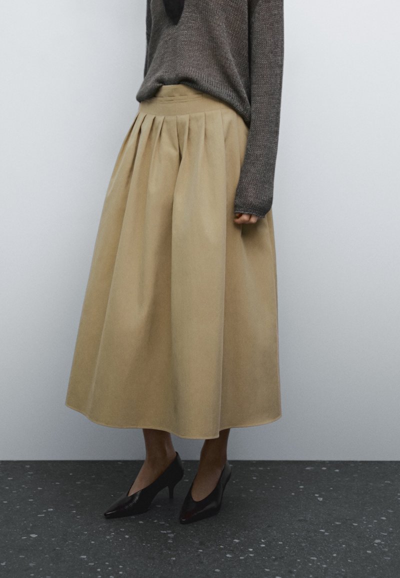 Personne portant une jupe midi plissée beige, un pull à manches longues gris foncé et des chaussures noires à talons hauts, debout sur un sol sombre tacheté.