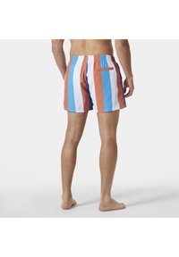 Mężczyzna w wielokolorowych pionowych kąpielówkach w niebieskie, białe i pomarańczowe paski, stojący boso na białym tle.