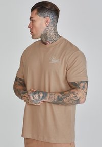 Tan short-sleeve t-shirt with a round neck, featuring a small white logo on the chest and smooth, soft fabric texture.