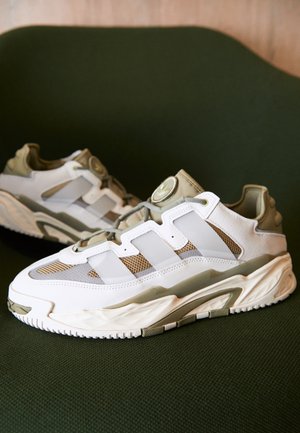 Białe i beżowe buty sportowe Adidas z siateczką i skórzanymi detalami, umieszczone na ciemnozielonej teksturowanej powierzchni, ustawione pod kątem, pokazując widok z boku i z przodu.