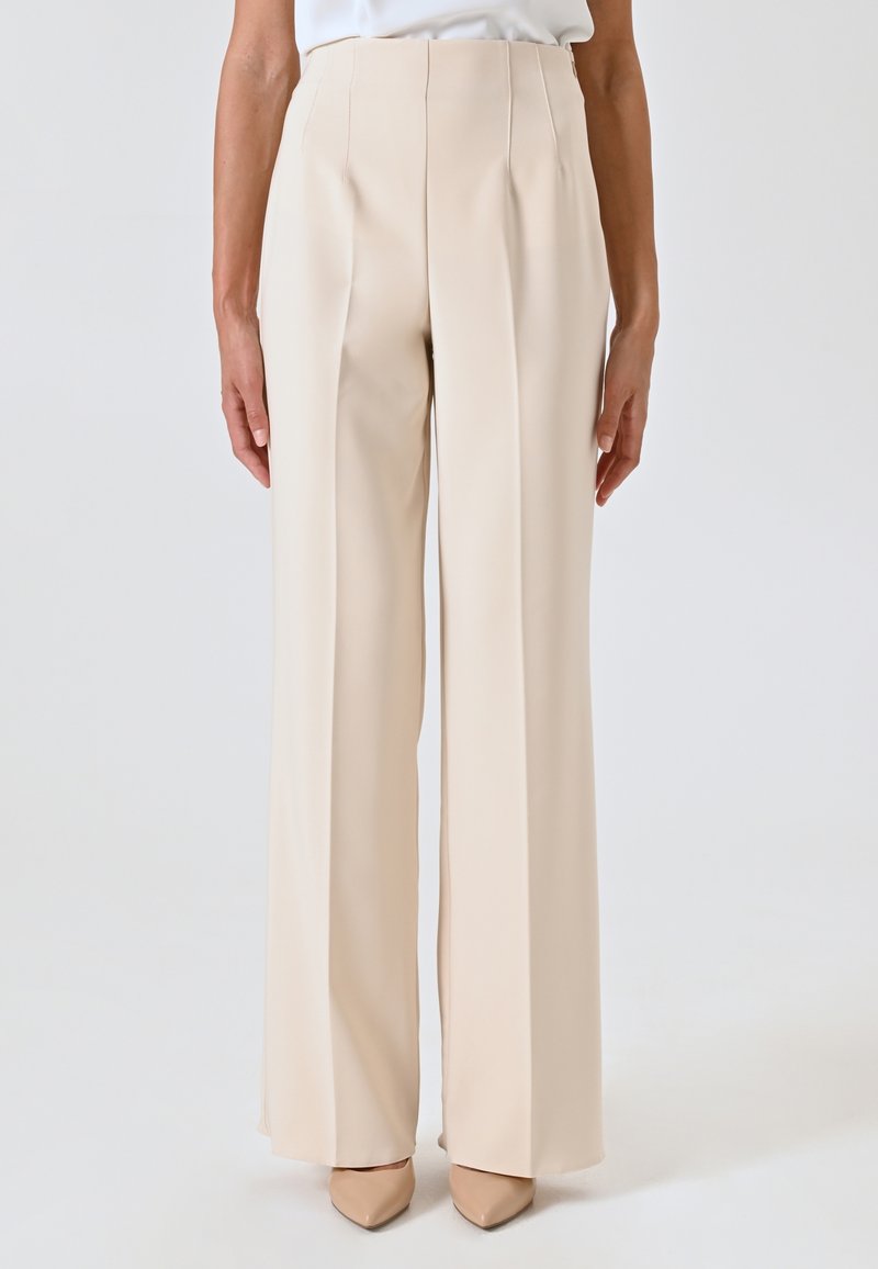 Beige, hoge taille broek met een rechte pijpdesign, gekenmerkt door scherpe plooien en een gladde textuur. Gecombineerd met nude topjes met een spitse neus.