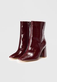Botas de tobillo burdeos hechas de cuero charol brillante. Presentan una puntera puntiaguda, un tacón cuadrado alto y un cierre de cremallera en la parte delantera.