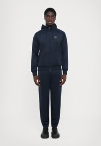 Survêtement navy comprenant un hoodie zippé avec cordons de serrage, des poches et un pantalon ajusté. Tissu texturé et poignets élastiques, associé à des baskets noires.