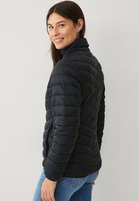Sort puffer-jakke med høj krave, med vandret quiltet design, lynlåslommer på siden og elastiske manchetter, båret over blå jeans.