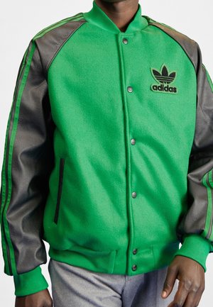 Homme portant une veste varsity Adidas verte et noire avec un logo sur la poitrine et un pantalon gris, debout devant un fond blanc.