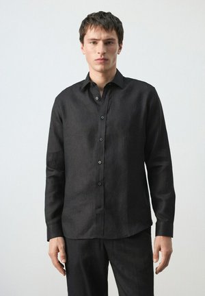 Joven con una camisa negra lisa abotonada y pantalones negros a juego, de pie frente a un fondo claro y sencillo.