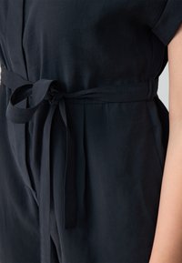 Marineblauwe jumpsuit met een cinched taille, voorzien van een stoffen riem en een zachte, gladde textuur. Korte mouwen en een losse pasvorm.