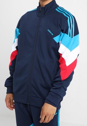 Osoba nosząca granatową kurtkę dresową Adidas z niebieskimi, białymi i czerwonymi pasami chevron na rękawach oraz jasnoniebieskimi paskami Adidas na ramionach.