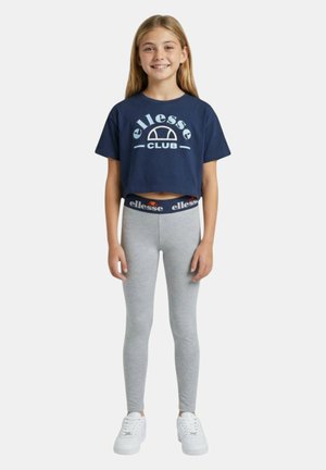 Ragazza sorridente in piedi che indossa un crop top blu navy con logo "ellesse CLUB", leggings grigi con vita "ellesse" e scarpe da ginnastica bianche su sfondo bianco.