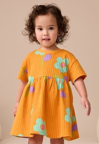 Vestido amarillo con textura de gofre y mangas cortas, con grandes flores verdes y círculos morados. Diseño acampanado y cintura elástica.