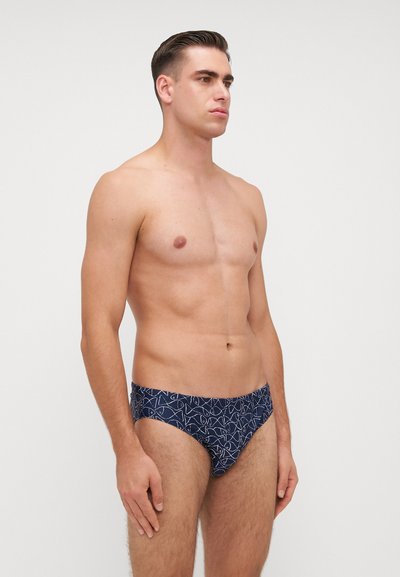 Navy zwemslips met een geometrisch wit patroon, gemaakt van rekbare stof, met een laag uitgesneden model en een gladde textuur.