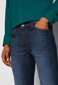 Jeans en denim bleu marine avec une coupe slim, des coutures jaunes contrastantes, un design à cinq poches et une fermeture à bouton et zip visible.