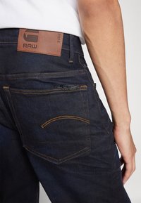 Mørkeblå denimjeans med orange syninger, et lædermærke mærket "RAW" og et klassisk fem-lomme design.