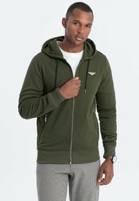 Sudadera con cremallera de color verde oliva con tejido texturizado, capucha ajustable, bolsillos frontales y detalle del logo en el pecho. Pantalones grises con un patrón.