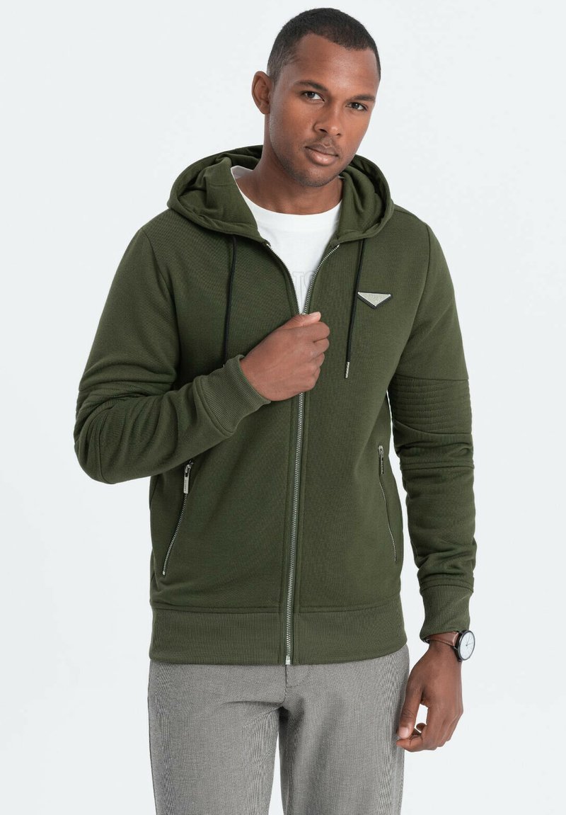 Sudadera con cremallera de color verde oliva con tejido texturizado, capucha ajustable, bolsillos frontales y detalle del logo en el pecho. Pantalones grises con un patrón.