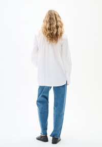 Weißes Langarmhemd mit gerafftem Rücken, kombiniert mit blauen Denim-Hosen mit weitem Bein und schwarzen Slippern, von hinten betrachtet.