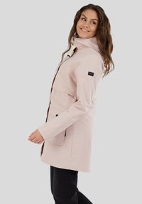Manteau imperméable rose clair avec un col haut, une fermeture éclair sur le devant et des poches latérales. Présente des accessoires noirs et une taille cintrée pour un look ajusté.