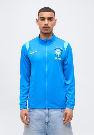 Fiatal férfi, kék Brazil labdarúgó csapat dzsekit visel, rajta a CBF logóval és sárga Nike szárnyalattal, egyszerű fehér háttér előtt áll.