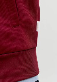 Κοντινή λήψη υφάσματος μπορντό που δείχνει ριγωτό μανσέτα και μια μικρή λευκή ετικέτα με πλαγιογραμμένο κείμενο προσαρτημένη στη ραφή.