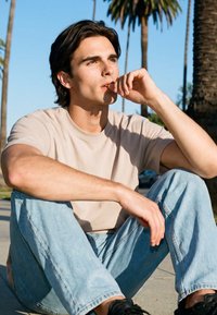 Mladi moški v bež majici in svetlih kavbojkah sedi na odprtem na pločniku, opira brado na roko, v ozadju so palme in modro nebo.