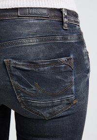 Primer plano del bolsillo trasero de unos vaqueros de mezclilla azul oscuro con textura descolorida y costuras decorativas, usados con una camiseta de punto blanca.