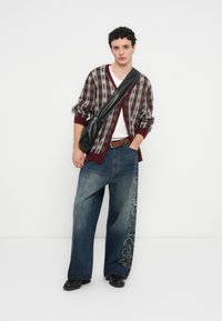 Prugasti kardigan u boji tamnocrvene i sive, bijela majica ispod, široke traperice plave boje s vezenim detaljima, crna torba i čizme.