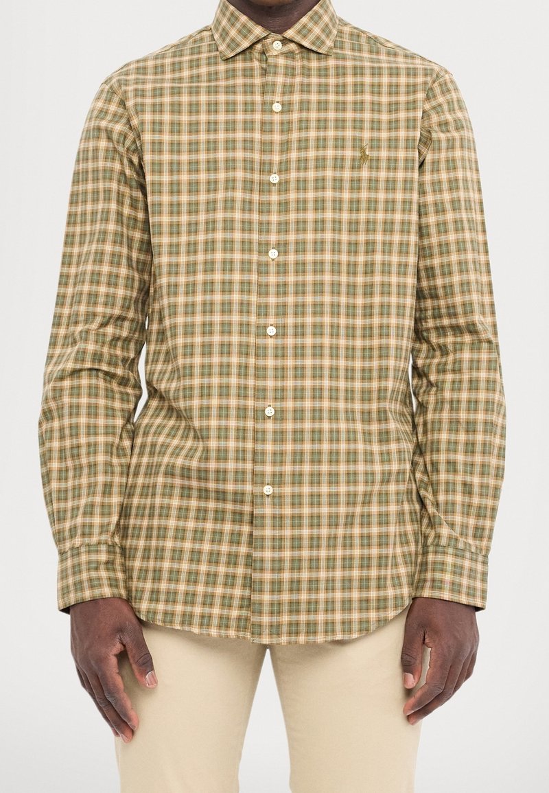 Man klädd i en långärmad skjorta med grön, beige och vit rutigt mönster samt beiga byxor, stående med armarna avslappnat längs sidorna.