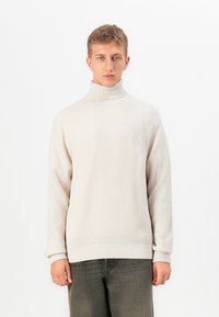 Young man wearing a cream turtleneck sweater and dark gray pants, standing against a plain white background.